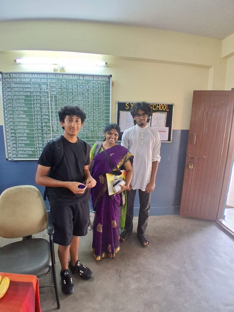 Two Finance For All volunteers with a teacher at Sri Yogeshwarananda English Primary & High School in Bangalore, standing in front of the school's staff board